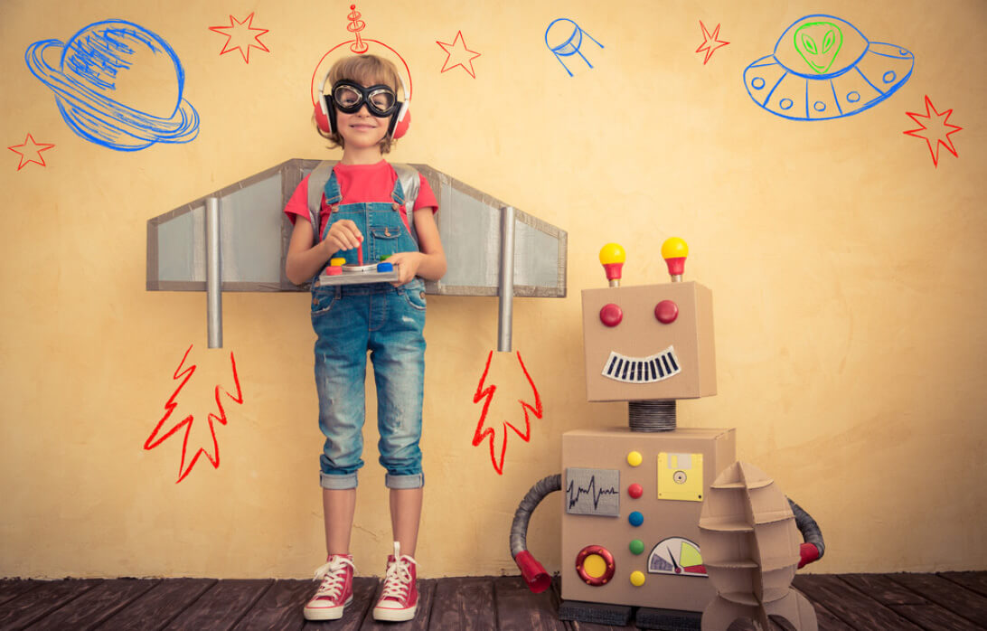 Natural Pharmaceuticals „OmegaMarine Junior+“ Vaikas su skraidančia mašina ir robotu iš kartono, fonas su kosmoso piešiniais žaidžia kūrybiškai.