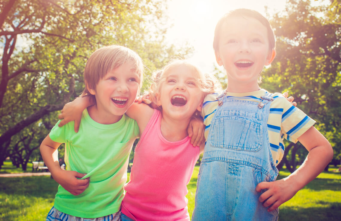 Natural Pharmaceuticals „OmegaMarine Junior+“ Trys linksmai šypsantys vaikai žaliame parke saulėtą dieną.