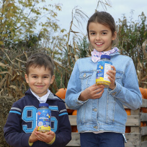 Natural Pharmaceuticals „OmegaMarine Junior+“ Vaikai lauke šypsosi ir laiko vitaminų buteliukus rudens daržo fone.