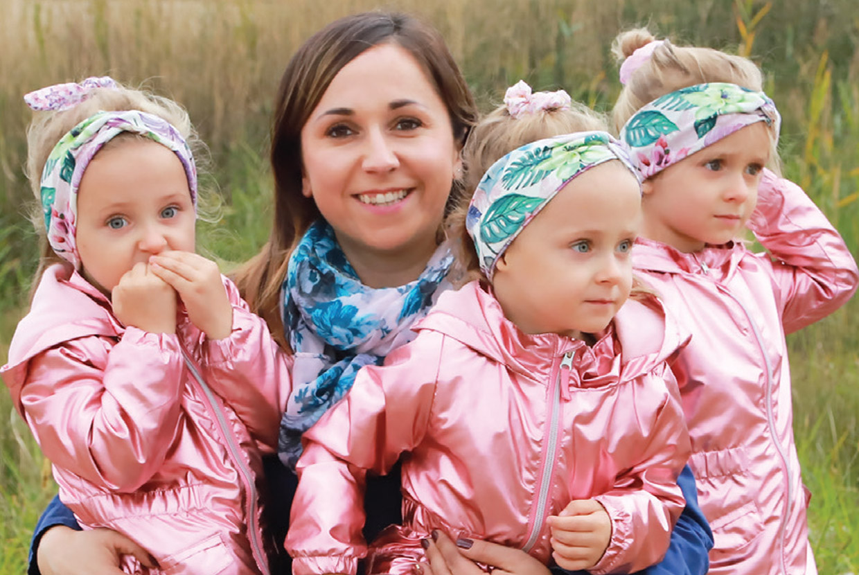 Natural Pharmaceuticals „OmegaMarine Junior+“ Motina apkabina tris mažas mergaites, apsirengusias identiškais rožiniais kombinezonais ir plaukų juostelėmis.