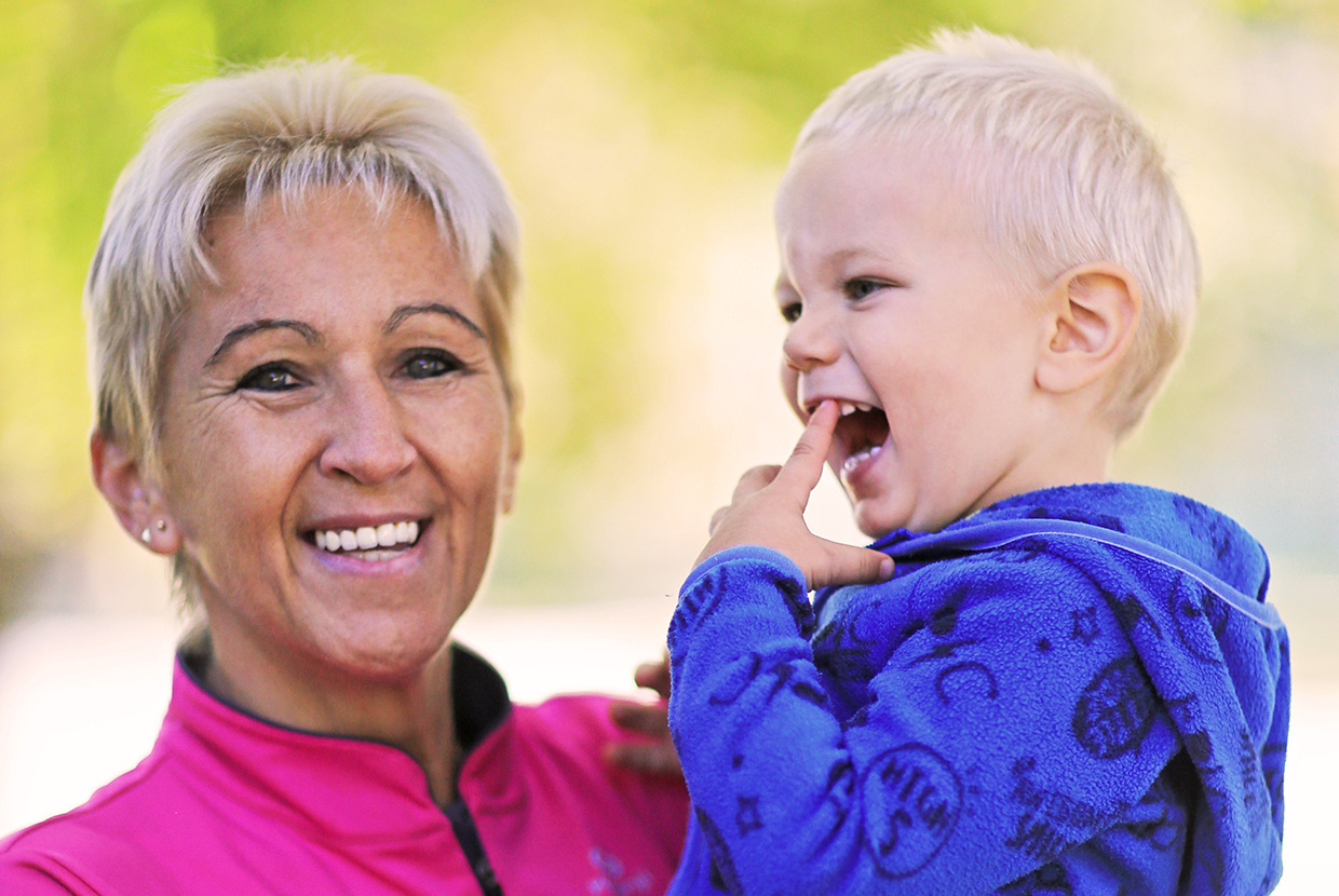 Natural Pharmaceuticals „OmegaMarine Junior+“ Šypsanti moteris laiko juokiančią mažametį vaiką mėlynoje striukėje lauke šviesiame fone.